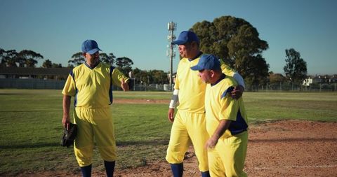 Baseball teammates walking on diamond helping hunched player in yellow and blue uniforms