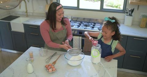Mother and daughter baking together on marble countertop, sifting flour and smiling