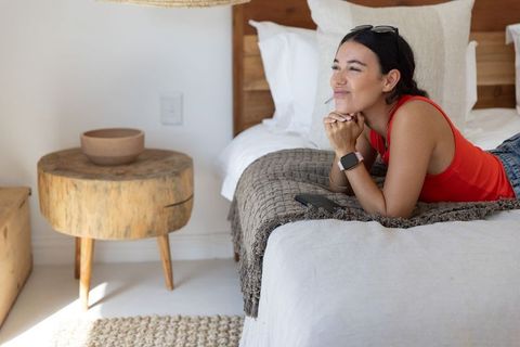Young woman lying on bed relaxing and daydreaming in cozy minimalist bedroom basking in morning ligh
