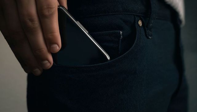 Hand sliding sleek smartphone into front pocket of dark blue denim jeans, closeup