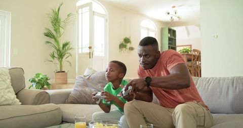 African American Father and Son Playing Video Games on Sofa in Sunlit Living Room