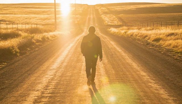 Solitary walker strolling down sunlit gravel road at golden hour with warm lens flare