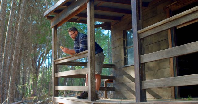 This image depicts a man standing on the porch of a rustic wooden cabin in the middle of a forest. He is focusing on what appears to be a smartphone in his hands. The surrounding trees and natural environment suggest a peaceful and isolated rural retreat. This can be used for themes related to nature retreats, solitude, leisure activities, digital detox, and outdoor hobbies.