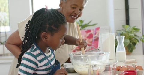 Grandmother guiding granddaughter stirring batter while baking together in bright kitchen