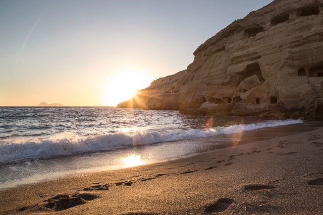 Photo features a sunrise over a rocky beach with gentle ocean waves lapping against the sandy shore. Footprints in the sand add a serene, tranquil touch, with tall cliffs framing the scene. Ideal for use in travel brochures, holiday advertising, or inspirational content highlighting the beauty of nature. Perfect for emphasizing themes of serenity, early mornings, and natural landscapes.