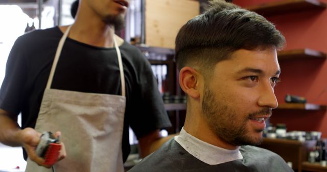 Man sits in barbershop chair while barber in apron provides haircut. Ideal for men's grooming services, hairstyling tutorials or advertisements for barbershops.