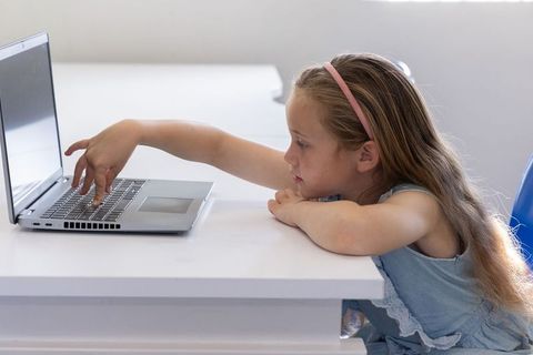 Young girl reaching for laptop on white desk in bright minimalist room for remote learning