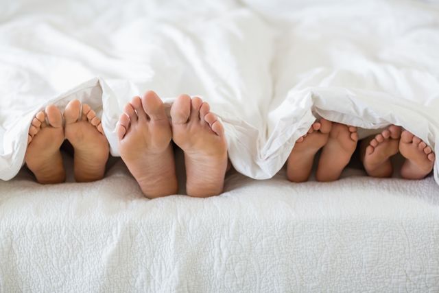 Feet of parents and children peeking out from under a white blanket in bed. Perfect for illustrating family bonding, cozy home environments, parenting moments, and concepts of relaxation and comfort.