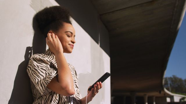 Young biracial woman enjoys music through her smartphone while basking in the sun at an urban park. Dressed in casual attire, she exudes tranquility and joy. Suitable for illustrating lifestyle posts, ads for mobile devices or audio products, and content about leisure and well-being in urban settings.