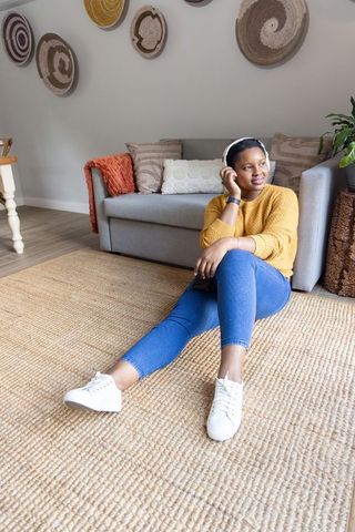African american woman relaxing on rug with headphones, smartphone and cozy mustard sweater