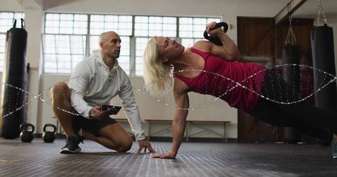Female athlete performing kettlebell side plank while trainer coaching with tablet