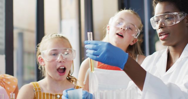 African American female scientist wearing lab coat and safety goggles teaching school children basic chemistry experiment. Two excited children wearing protective goggles and summer clothes are observing her. Ideal for educational content, school science program promotions, STEM campaigns, and diversity in science initiatives.