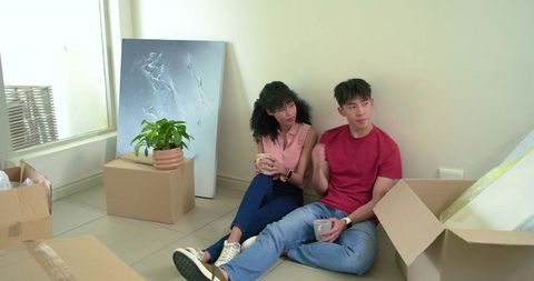 Diverse young couple unpacking in new apartment, sitting on floor sipping coffee, relaxing