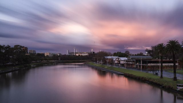 Serene river landscape captured during sunset, showcasing dramatic clouds and pink sky reflecting on the water. Long exposure adds a sense of calm and tranquility to the scene. Perfect for travel ads, urban development presentations, nature blogs, and relaxation-themed visuals.
