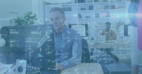 Man typing at desktop in modern open-plan office with glass reflections, monitors and coworking