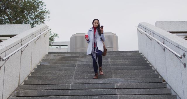 Asian woman walking down the stairs talking on smartphone and holding takeaway coffee - Download Free Stock Photos Pikwizard.com