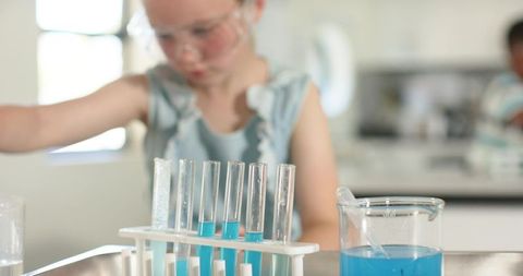 Young student wearing safety goggles mixing blue liquids in test tubes in classroom lab