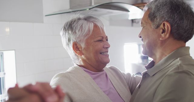 Senior couple dancing in kitchen, sharing a joyful and intimate moment. Both are smiling warmly, enjoying their time together. Ideal for use in advertisements for retirement communities, healthcare services, or senior lifestyle publications. Conveys themes of love, happiness, and an active, fulfilling lifestyle for older adults.
