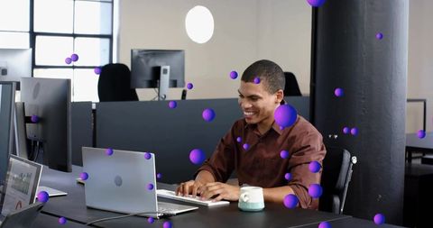 Young man smiling typing at laptop in modern open-plan office with purple bokeh overlays