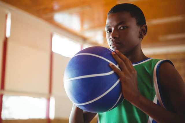Young male basketball player standing in an indoor court, holding a blue basketball and looking confident. Ideal for use in sports-related content, youth athletic programs, basketball training materials, and motivational posters.