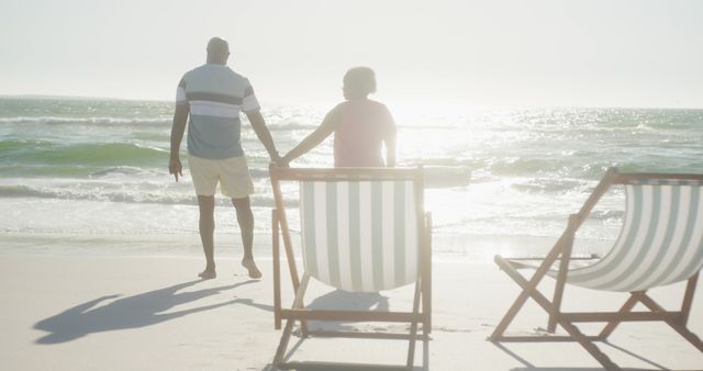 Happy senior african american couple holding hands and walking on sunny beach at sunset, copy space. Retirement, summer, vacations, togetherness and senior lifestyle, unaltered.