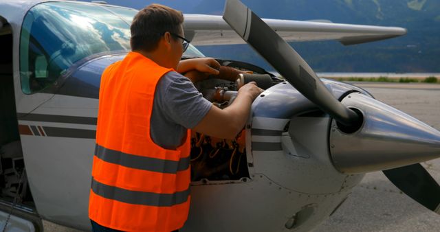 Mechanic wearing safety vest examining and repairing engine of light aircraft on an airfield. This can be used for topics related to aviation maintenance, airplane inspections, aviation safety, technical jobs, airfield operations.