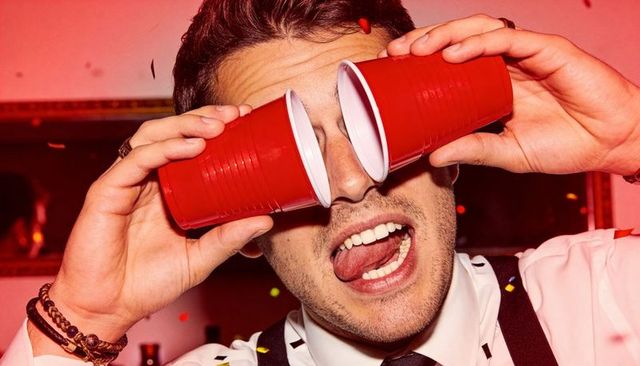 Playful man holding red party cups over eyes sticking out tongue closeup with confetti