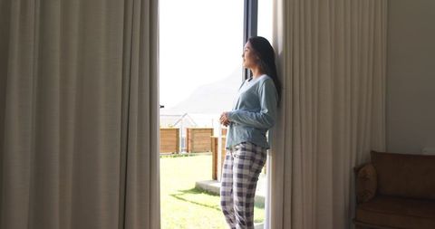 Mid-adult asian woman standing at patio door wearing plaid pajamas and enjoying morning sunlight