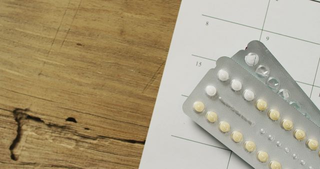 Blister packs of birth control pills lie atop a wooden table next to a calendar, indicating a method of family planning and scheduling. This image is useful for topics related to family planning, contraceptives, hormone therapy, and healthcare scheduling.