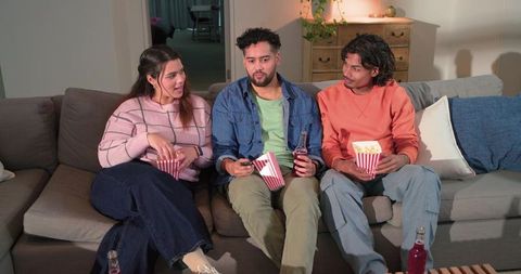 Hispanic friends watching movie on sofa, sharing popcorn and drinks in cozy living room