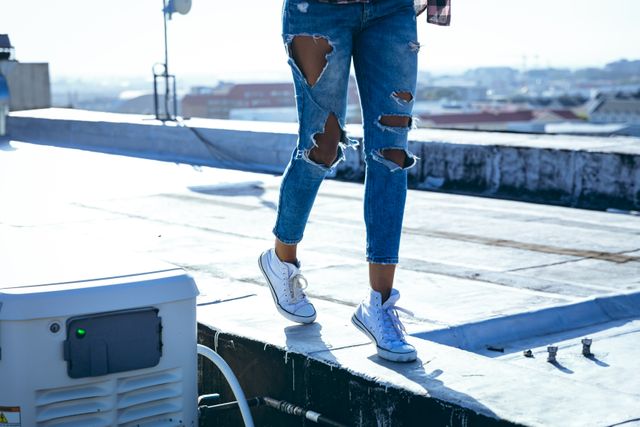 Front view low section of a hip young biracial woman wearing jeans and white trainers, walking on an urban rooftop with buildings in the background.