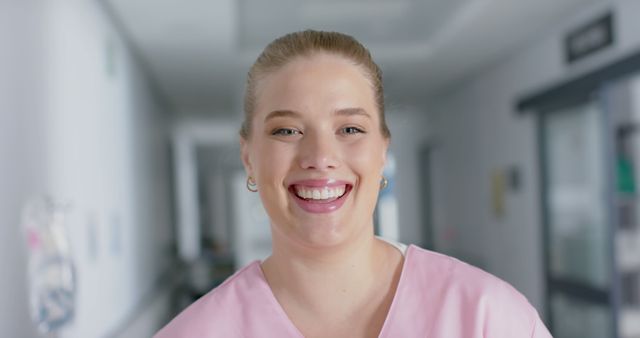 Nurse in pink scrubs smiling warmly while standing in a hospital corridor. Ideal for representing healthcare services, nurse appreciation, patient care, and hospital environments. Useful for medical websites, promotional materials, and health-related social media posts.