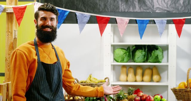 Smiling vendor with a beard wearing an apron standing in front of a market stall full of fresh fruits and vegetables. The stall is decorated with flags. Ideal image for promoting farmers' markets, organic food, local produce, and healthy eating.