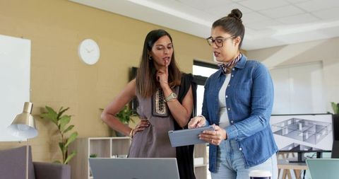 Diverse women collaborating on tablet in modern office with laptop and floorplan on monitor
