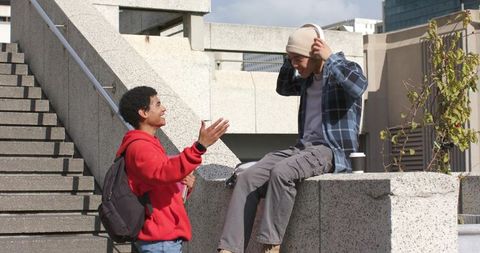 Young men chatting on urban steps one adjusting headphones with coffee