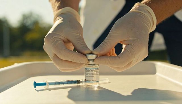 Preparing vaccine vial outdoors with gloved hands and syringe on tray for inoculation