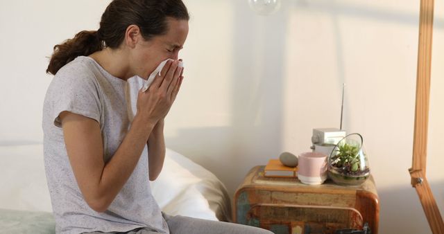 Woman sitting on bed sneezing into tissue, indicating possible cold, flu, or allergies. Bed is adjacent with nightstand holding books, potted plant, and other small items. Useful for healthcare, medical, home setting, allergy treatment, and wellness themes. Image depicts personal space and disease prevention measures.