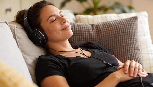 Contented young woman relaxing on couch with wired headphones and cushions at home