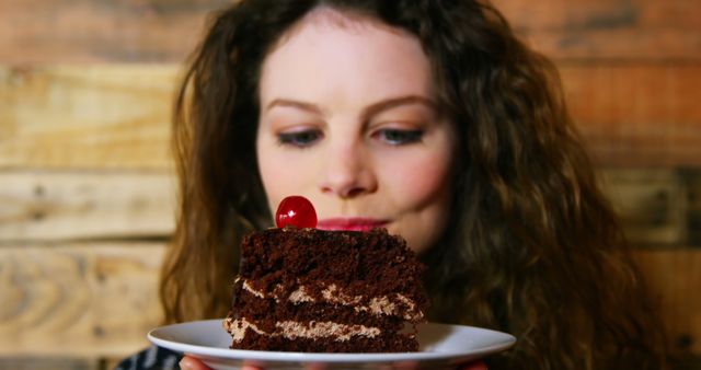 Curly-haired woman admiring a slice of chocolate cake topped with a cherry. Perfect for illustrating dessert enjoyment, food experiences, or bakery promotions. Rustic wooden background adds a cozy atmosphere. Ideal for blogs, culinary websites, and advertisements related to sweets.