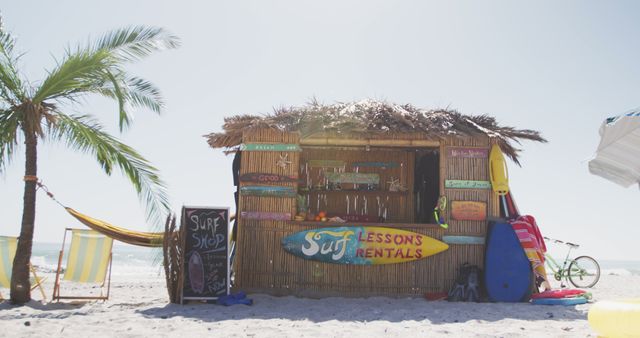 Tropical surf shop hut on a sunny beach. Palm tree provides shade, while surfboards, signs, and colorful equipment are displayed in and around the hut. Ideal for travel, tourism, and adventure-themed projects, this image highlights the relaxed atmosphere of beach life and surf culture.