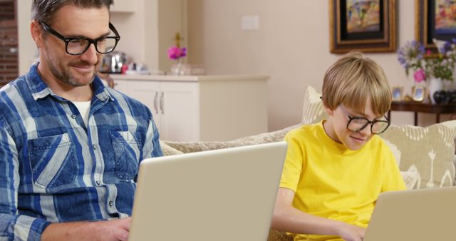 Father and son are sitting on the couch with laptops, working side by side. Both are wearing glasses, with the father in a blue plaid shirt and the son in a yellow shirt. Framed pictures and flowers are in the background, giving a homely atmosphere. This can be used to represent family activities, remote work, learning, bonding, or technology use at home.
