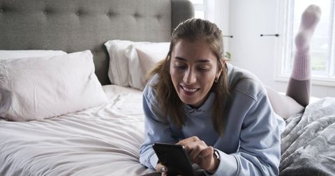 Young woman lying on bed using smartphone, relaxing in pastel bedroom, cozy lifestyle
