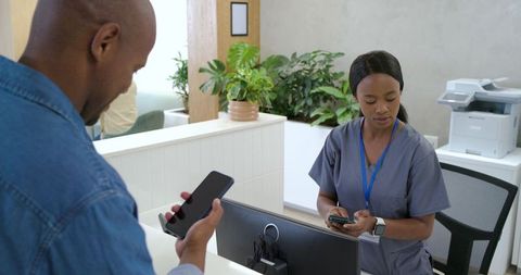 African american receptionist checking phone with patient at clinic front desk, healthcare
