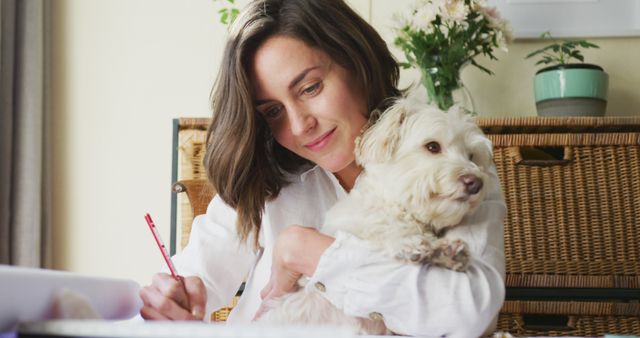 Young woman working at home with her dog on her lap, showing warmth and companionship. She is writing with a pencil while happy and relaxed. Perfect for use in articles on work-life balance, love for pets, remote working, and creating a home office environment.