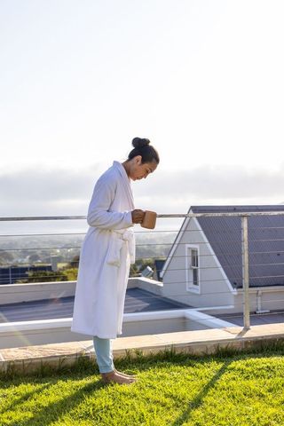 Rooftop morning ritual: woman in white robe holding ceramic mug, barefoot on grass