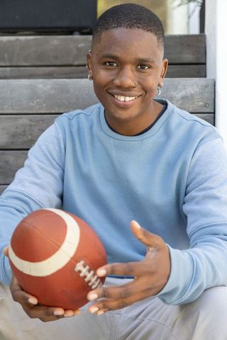 Smiling african american teen holding football on porch wearing crewneck and nose ring