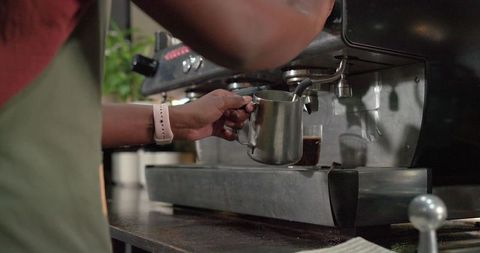 African american barista frothing milk with stainless steel pitcher at espresso machine