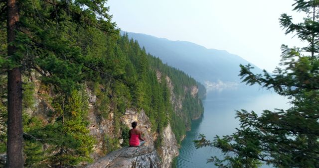 Woman enjoying serenity perched on a cliff, around surrounded pine trees, stunning lake view. Perfect for travel, adventure, nature themes, relaxation concepts in ads, blogs, publications.