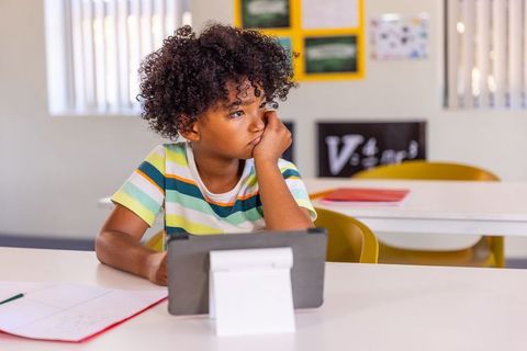 Pensive african american boy sitting at desk gazing at tablet in bright classroom