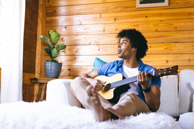 African American man sitting on a white couch in a cozy log cabin, playing an acoustic guitar. The wooden interior and natural light create a warm and inviting atmosphere. Ideal for use in lifestyle blogs, music-related content, or advertisements promoting relaxation and leisure activities.